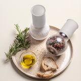 Two Tupperware spice grinders with salt and peppercorns on a tray with olive oil, bread, and rosemary.
