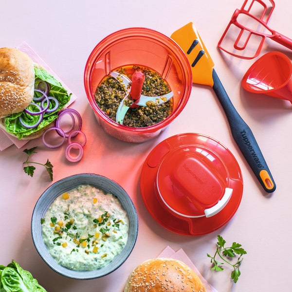 Red manual food chopper with fresh herbs, dip, spatula, and burger ingredients on kitchen counter